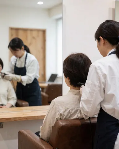A stylist cuts a client's hair in a salon chair, reflected in a mirror against white walls and a wooden door.