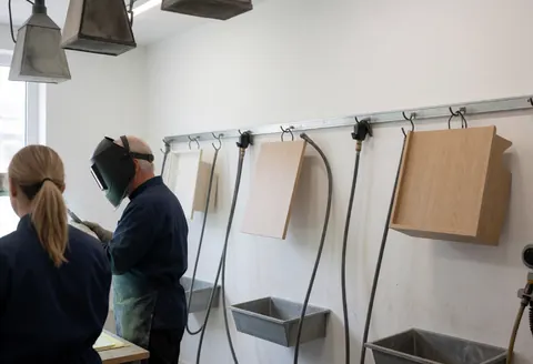 Two workers wearing protective gear restore hanging furniture in a workshop with white walls and metal containment boxes.