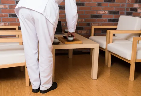 Yoga instructor in white uniform serving tea on a wooden tray at a table in a lounge with brick walls and chairs