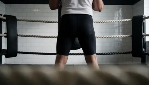 Person standing with back to camera in a boxing ring with white tiled walls, suitable for gym or boxing academy website hero image