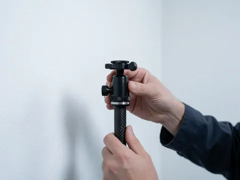 Close-up of a hand tightening the locking mechanism on a black carbon fiber monopod against a white wall with cool professional lighting