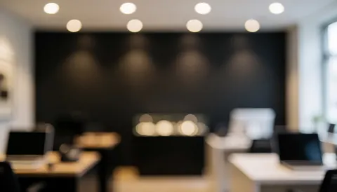 Dark wall office background with desks, laptops, and soft ceiling light bokeh, suitable for text overlay