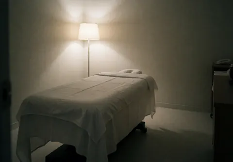 Massage bed with white sheets in a minimalist room with neutral walls and soft floor lamp lighting