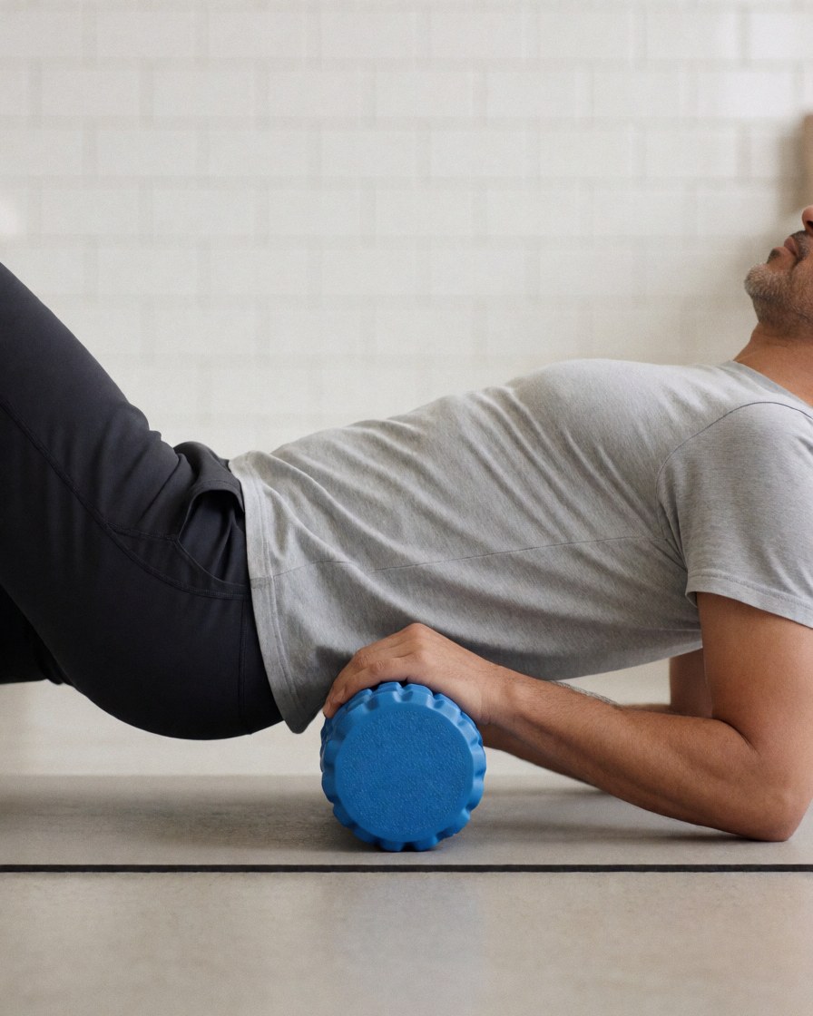 Man using blue foam roller on yoga mat in gym with white tiled walls