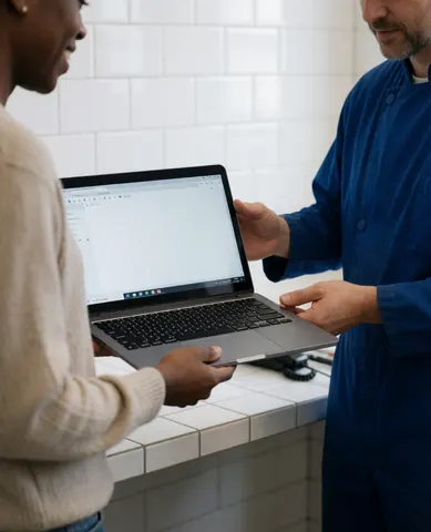 A technician in a blue uniform hands a powered-on laptop to a customer in a workshop with white tiled walls.