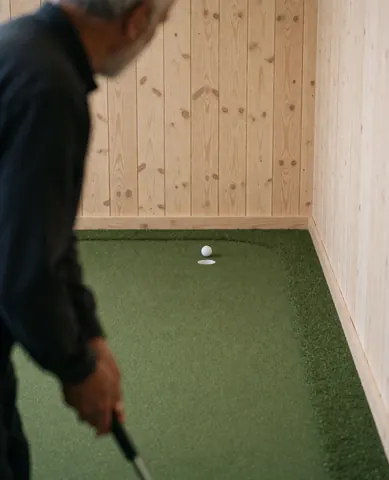A golfer from behind preparing to putt with a putter, golf ball, and hole visible on a wooden-walled indoor simulator