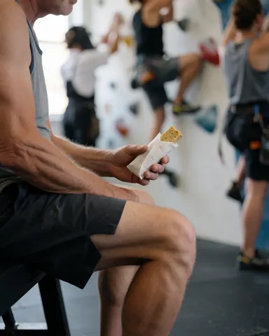 A climber rests on a bench holding a wrapped protein bar while other climbers scale the wall in the background.