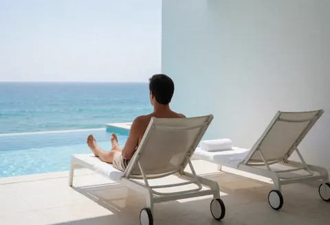 A man rests on a white lounge chair beside an infinity pool merging with the ocean, featuring white walls and an empty adjacent chair.