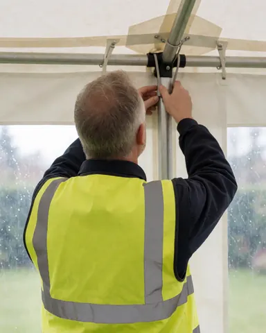 Worker in reflective vest adjusting metal canopy frame from behind during outdoor banquet setup