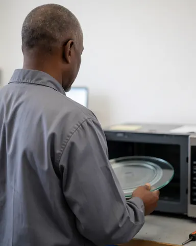 Technician placing a new glass turntable into an open microwave oven in a workshop with white walls.