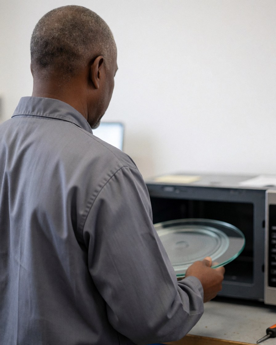 Technician placing a new glass turntable into an open microwave oven in a workshop with white walls.