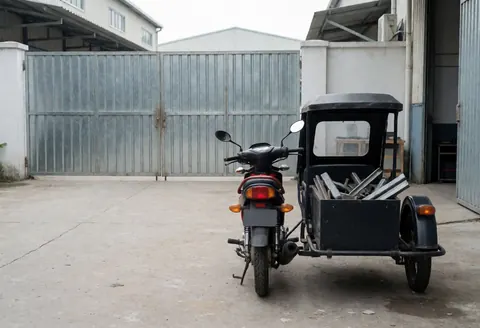 Motorcycle taxi with sidecar loaded with metal parked in front of industrial workshop door on concrete ground under cloudy sky