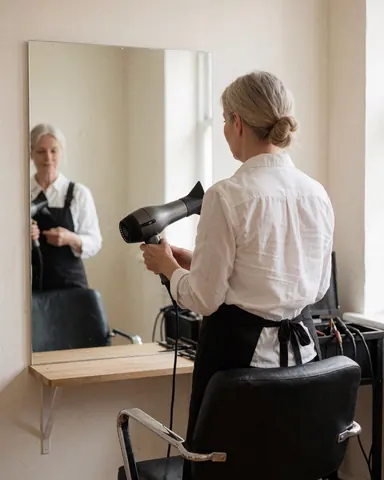 A hair stylist using a hair dryer in a salon with a mirror and chair, light walls, professional scene