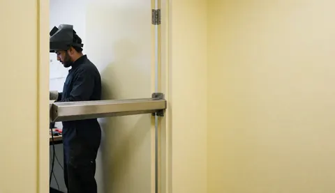 Security technician installing a magnetic bar on an office door with yellow walls and visible tools during an alarm repair service.