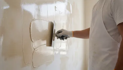 Painter applying Venetian stucco with trowel on white wall, wearing gloves and apron, glossy finish