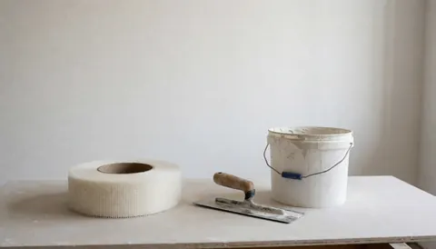 A roll of fiberglass mesh tape and a trowel resting on a sheet of drywall board for plastering and wall finishing work