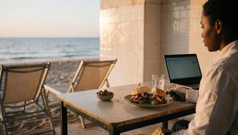 Person working on laptop at beach table with octopus sandwich, olives, and ocean view