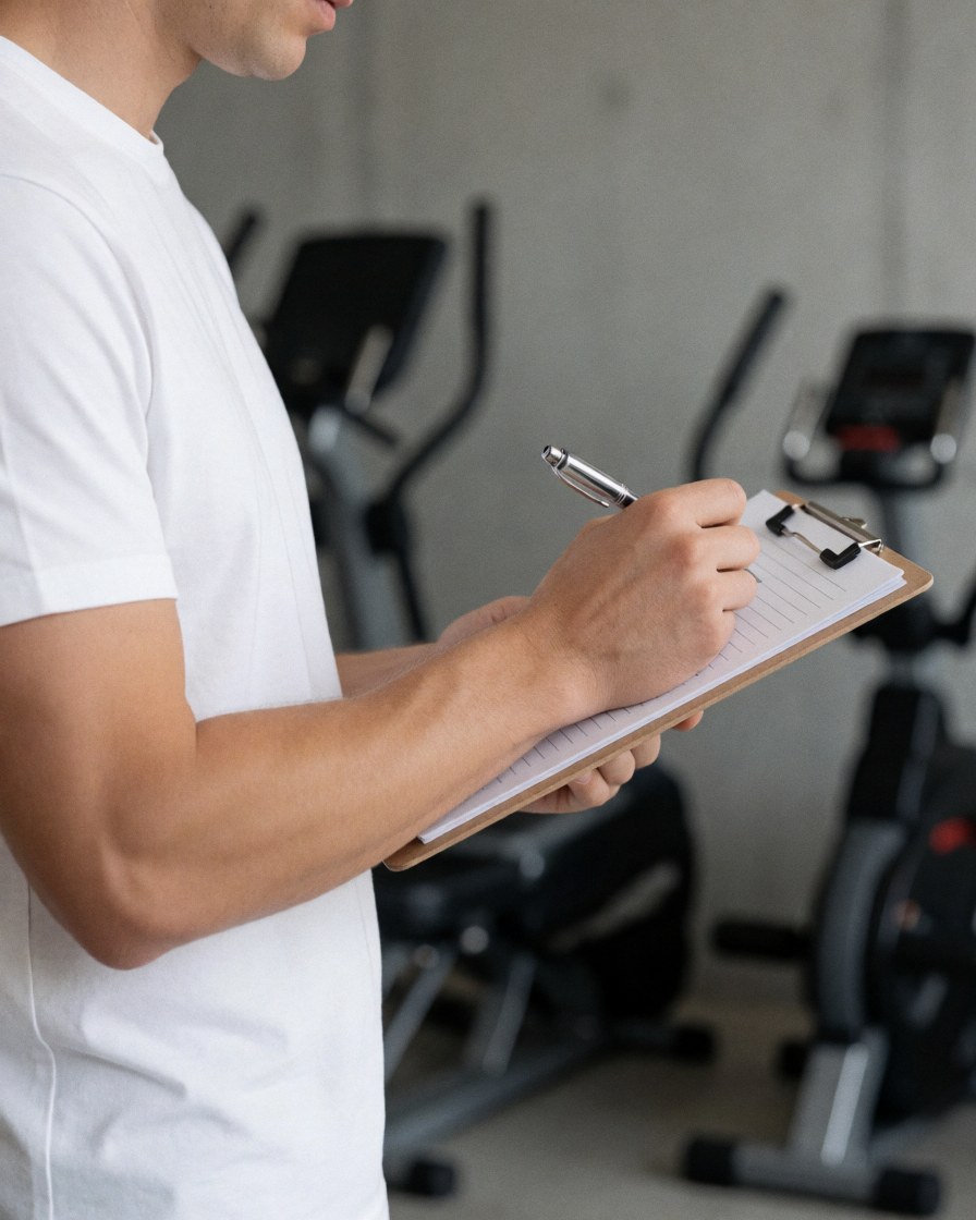 Personal trainer writing notes on a clipboard with gym equipment and concrete wall in the background