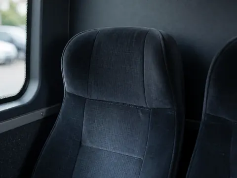 Empty bus seat featuring dark fabric and visible stitching against gray walls with ambient blue lighting, suitable for public transport contexts.