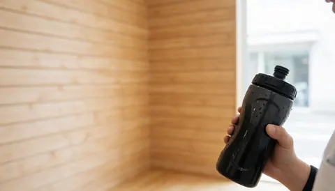 Close-up of a hand gripping a water bottle with condensation drops in a climbing gym featuring light wood walls and natural lighting