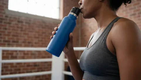 Woman drinking water from a blue bottle while standing in a boxing ring with a brick wall background and natural lighting