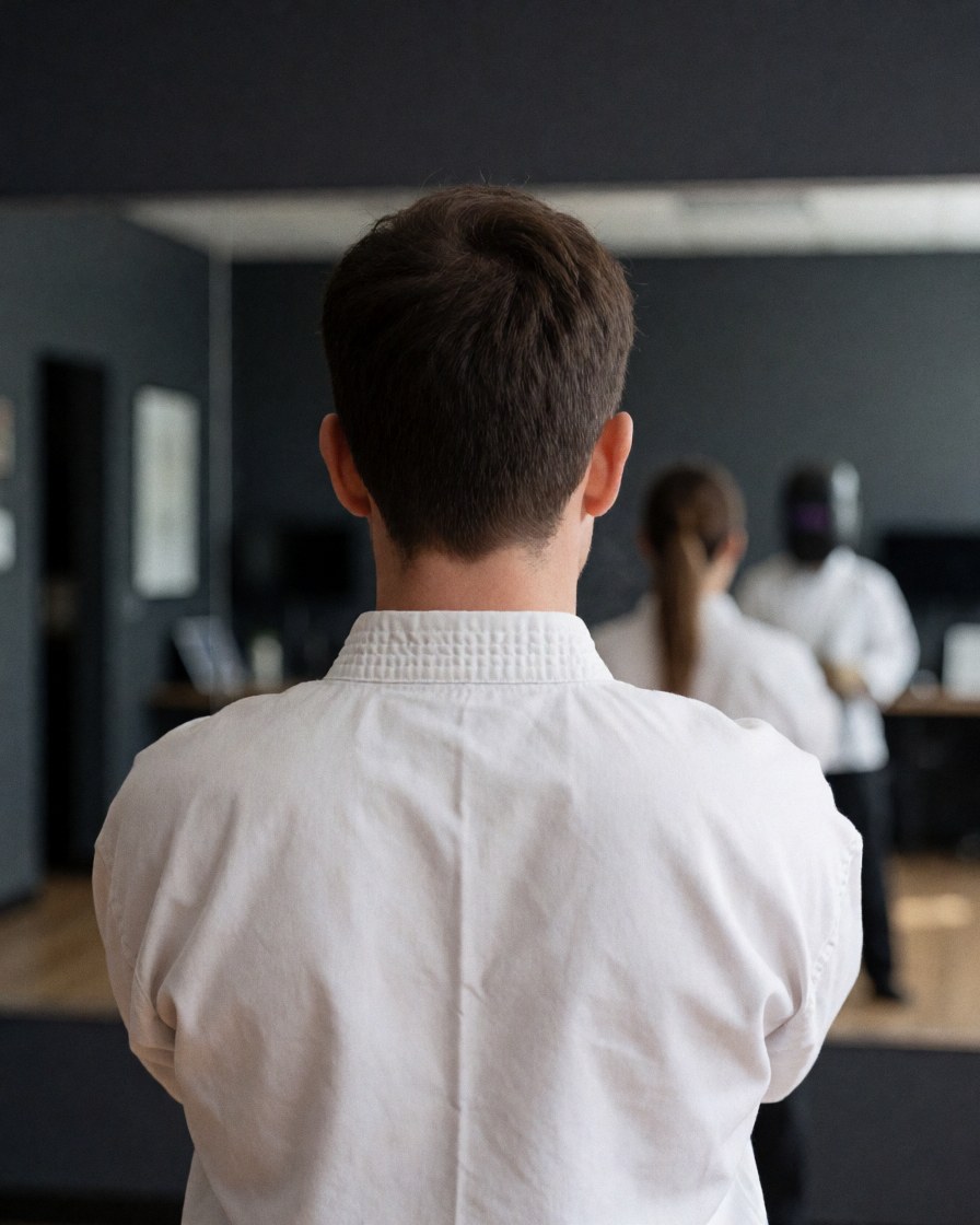 Back view of a martial arts student wearing a white dobok in a dojo with mirrors and gray walls