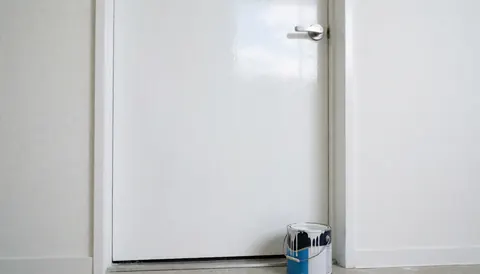 A freshly painted white door with a synthetic paint bucket on the threshold, white walls, and sky reflection, ideal for house painters.