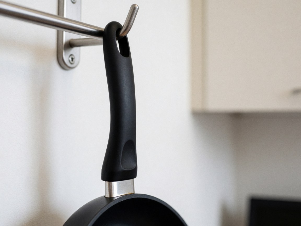 Close-up of a black frying pan handle suspended from a metal holder in a kitchen with white walls and blurred cabinets.