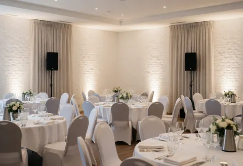 Elegant event hall featuring round tables, white chair covers, professional speakers, white brick walls, and soft ambient lighting.