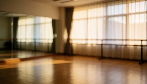 Empty dance studio featuring wooden flooring, wall mirrors, and a ballet barre, illuminated by warm natural light from curtained windows.