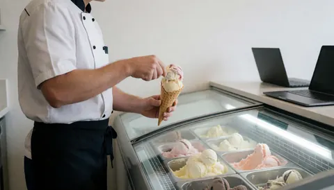 Ice cream vendor serving a fresh cone from a cooler, wearing a white and black uniform against a clean modern background.