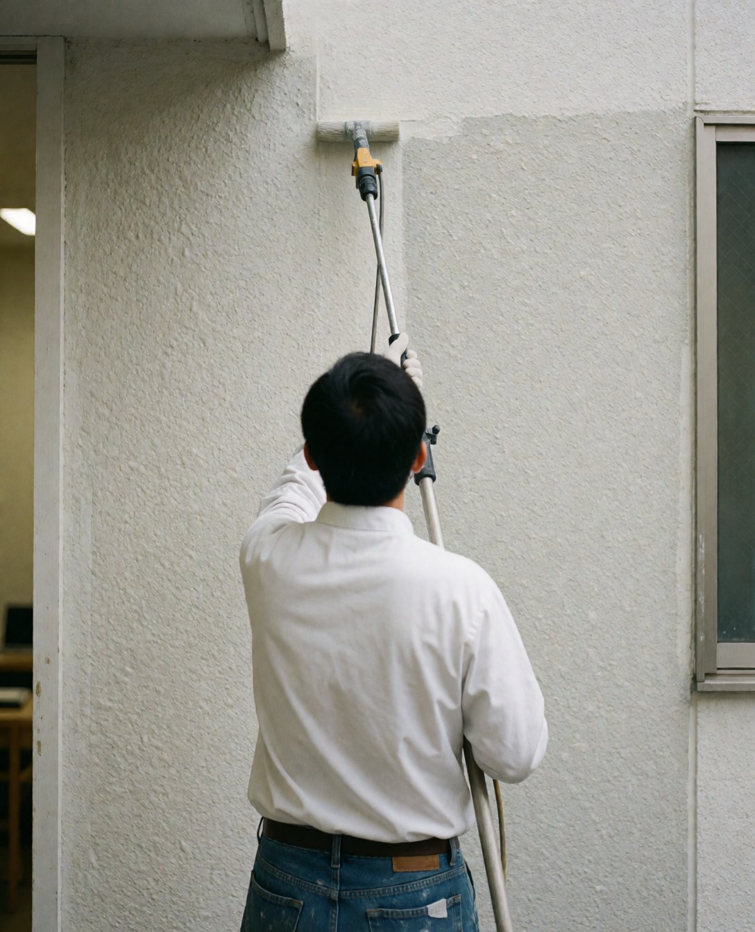 A house painter applies sealant to a textured exterior wall using a roller extension, viewed from behind.