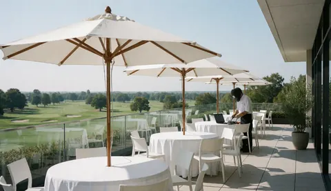 Terrace with round tables and white umbrellas overlooking the golf course, person working at a table