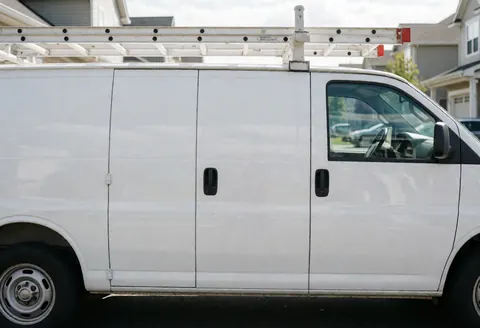 White service van equipped with a ladder parked on a residential street, ready for heating repairs with reflections visible in the windows.