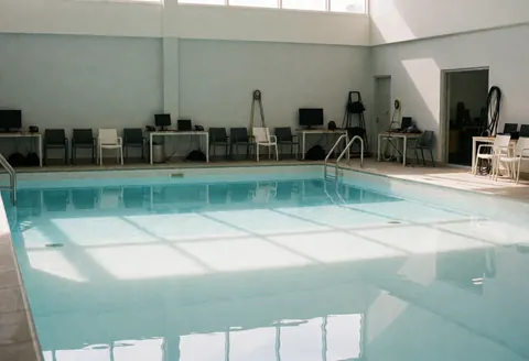 Empty public pool with calm water reflecting natural light, neatly arranged chairs and tables on the deck, and clean gray walls.