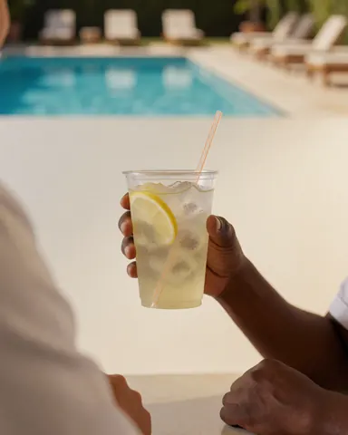 A hand holding a glass with lemon and ice in front of a pool with beach chairs, ideal for a water park.
