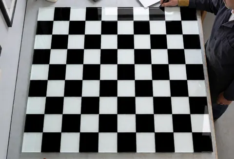 Black and white checkered decorative glass panel ready for installation in a workshop with gray walls and visible tools