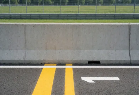Concrete wall with yellow stripes and white arrow on asphalt, grass and metal fence background