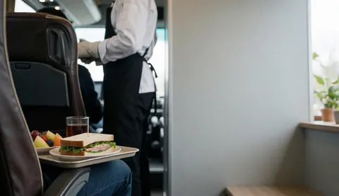A meal tray holding a sandwich, fresh fruit, and a beverage rests on a plush seat inside a tour bus with service staff visible in the background.