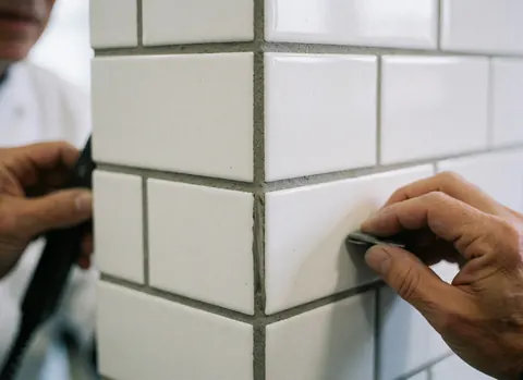 Close-up of a hand applying gray grout between white ceramic tiles using a sanding tool