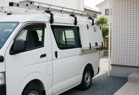 White commercial van equipped with ladder racks and a toolbox parked on gravel in front of a house with white tiled walls, ready for window installation.