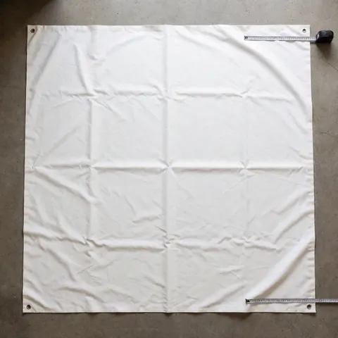 White heavy-duty awning cloth with metal corner hooks laid on concrete floor alongside a measuring tape for installation