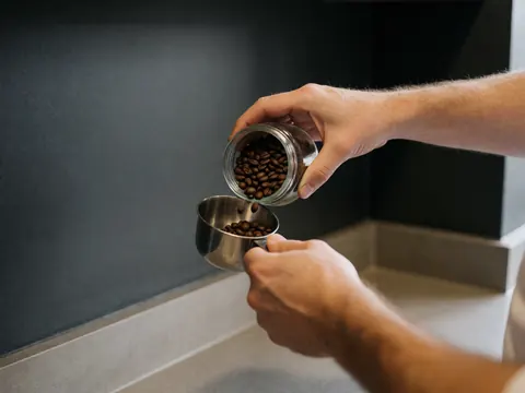 Whole coffee beans being poured from a glass jar into a metal spoon against a charcoal gray wall in an apartment hotel.