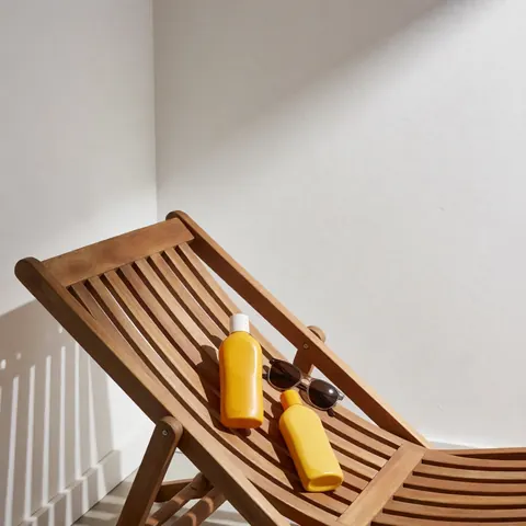 Wooden chairs with sunscreen bottles and sunglasses against white walls and sunlight shadows, ideal for a vacation rental.