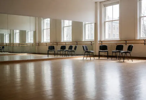 Empty theater rehearsal space featuring wall mirrors, ballet bars, black chairs, and wooden floors with natural light from windows.