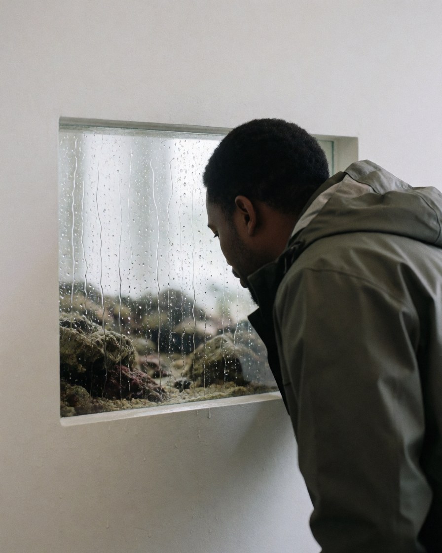 A visitor wearing a rain jacket looks at an aquarium with raindrops on the glass in a white-walled indoor space