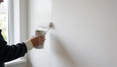 A professional painter uses a roller to apply white paint to a wall, leaving copy space, illuminated by natural light from a nearby window.