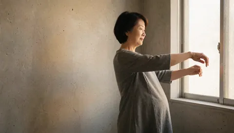 A pregnant woman performs gentle movements near a window with golden light and a concrete wall, suitable for a dance instructor.