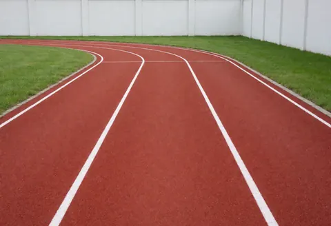 Empty curved red synthetic running track with white lane lines, green grass infield, and white walls in the background.