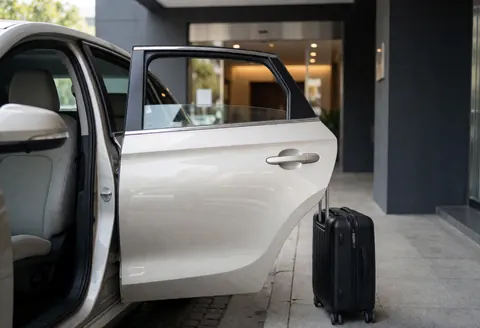 A modern black transport vehicle with its door open and a suitcase visible, parked outside a hotel ready for service.